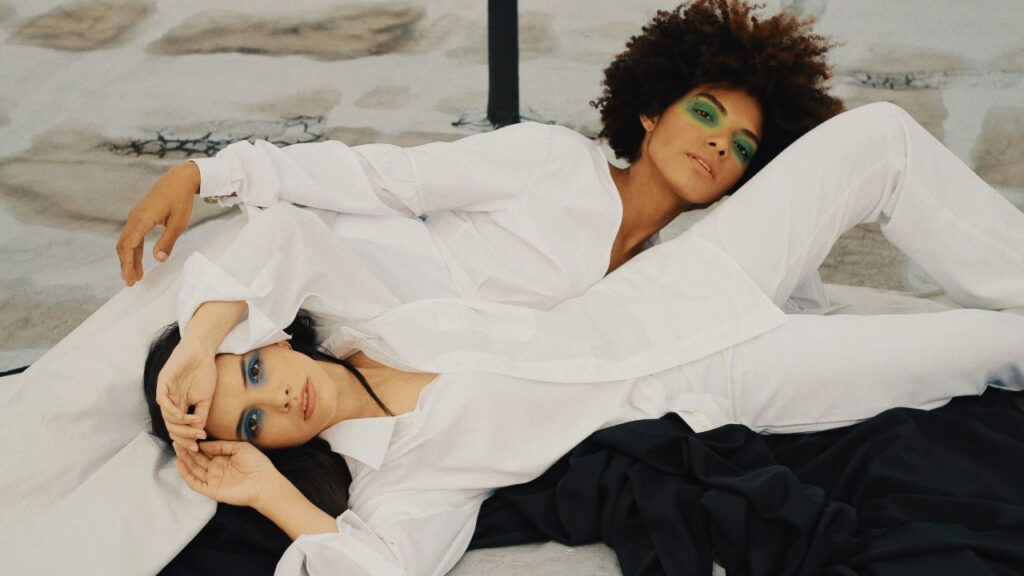 Dos personas con camisas blancas sobre una cama, imagen de moda minimalista en tonos neutros.